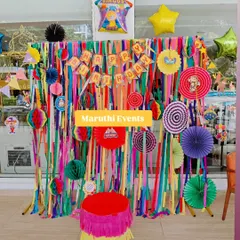 A colorful circus or carnival-themed party. The backdrop is made of vibrant streamers and paper fans, with a fringed plinth adding to the festive look.