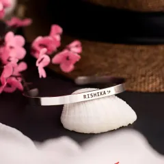 A silver cuff bracelet engraved with 'RISHIKA' and a small symbol, resting on a seashell. This product shot highlights the option to add small icons alongside a name.