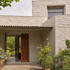 The entrance to Villa Uma in Alibaug features rough, textured walls and a simple corrugated roof. The design uses natural materials and deep overhangs to create a welcoming transition from the bright exterior to the cool, shaded interior.