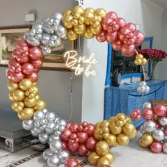A trendy and modern bachelorette party photo booth featuring a large circular balloon arch. The arch is made of metallic rose gold, gold, and silver balloons, with a "Bride to be" neon sign glowing in the center, perfect for a stylish home party.