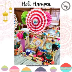 A festive spread of Holi hampers ready for gifting. These wooden trays are packed with snacks like chips and nachos, buttermilk, thandai, and colors, all decorated with colorful paper fans for a party-ready look.