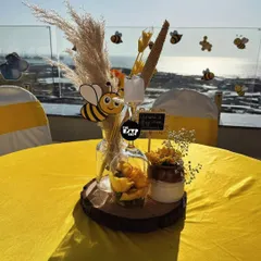A close-up of a tabletop centerpiece for our bee-themed baby shower. This arrangement sits on a rustic wood slice and includes pampas grass, a cute bee cutout, yellow flowers, and a small chalkboard sign, all set against a beautiful seaside view.