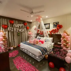 A full view of a romantic hotel room decoration for a birthday. The decor features a rose petal pathway, a bed canopy with lights, and 'I Love You' and 'Happy Birthday' foil balloons.