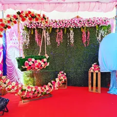 An outdoor naming ceremony setup with a floral crescent moon cradle. The backdrop features a green wall with hanging pink flowers, creating a fresh, garden-like atmosphere.