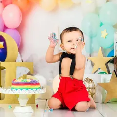 A baby boy with frosting on his hands smiles cheekily at the camera during his rainbow-themed cake smash. A picture of pure, uninhibited joy.