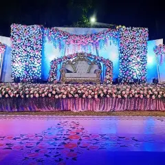 A wide-angle view of a spectacular floral reception stage at night. The setup is overflowing with pink and white flowers, with a cascading floral canopy and a beautifully decorated sofa for the couple, all lit to create a romantic and grand visual.