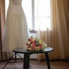 A simple, elegant shot of the wedding dress hanging by the window, with the bridal bouquet waiting on the table. A quiet moment of anticipation.