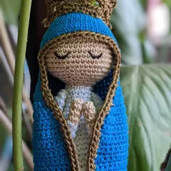 This serene Mother Mary doll is shown nestled amongst leaves, highlighting her peaceful presence. She wears a blue mantle and a golden crown.