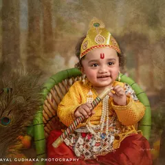 A happy one-year-old Krishna holds his flute, ready to charm the world. The setup includes a wicker chair and peacock feathers for a touch of rustic divinity.