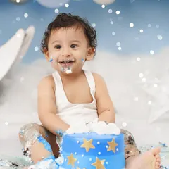A joyful baby boy with a frosting-covered smile enjoys his first birthday cake smash, set against a dreamy blue backdrop with moon and star props.