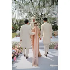 A walk to remember. The bride, escorted by her brothers, makes her way down a pristine white aisle lined with soft pastel flowers, heading towards a beautiful garden mandap for a serene and elegant farmhouse wedding ceremony.