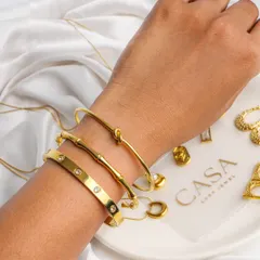 A close-up of gold bracelets on a model's hand, resting on a white plate with other jewellery pieces. The use of soft fabric and a human element creates a gentle, luxurious feel.