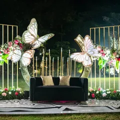 A whimsical and elegant photo booth for a Sangeet night. This setup includes a plush black sofa, large illuminated butterfly wings, and a dramatic black-and-white patterned stage.
