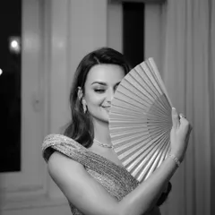 A playful and chic black and white portrait of the bride at her cocktail night, peeking over a fan with a smile.
