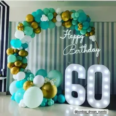 Celebrating a 60th birthday with a stylish ring backdrop. The teal, white, and gold balloon garland is paired with a "Happy Birthday" neon sign and large marquee numbers for a memorable setup.