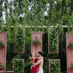 A couple in a loving embrace against a backdrop of lush greenery and hanging floral decor, a candid moment of romance.