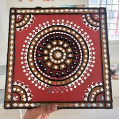 This square Lippan art piece features a traditional design in earthy red and brown tones, with intricate mirror work.