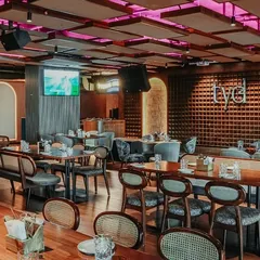 The main dining area combines various seating styles, from cane-back chairs to plush benches, all under a ceiling with acoustic panels and dynamic lighting.