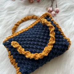 This handle bag is made with a dense, textured knot in a deep navy blue, accented with a ruffled tan border and a twisted handle. It's spacious and holds its shape well.