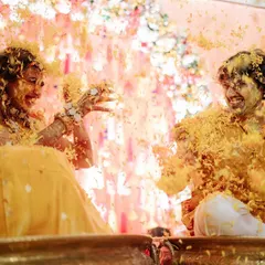 A beautiful, playful moment from a Haldi ceremony. The couple is covered in turmeric and flower petals, sharing a laugh in front of a delicate backdrop of pastel florals and hanging crystals.