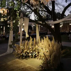 We created a dreamy, rustic photo corner using bunches of dried pampas grass, soft white drapes, and warm lighting. The chandelier hanging from the tree above adds a touch of elegance to the earthy setup.