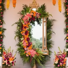 An asymmetrical floral arrangement beautifully frames this standing mirror. The design uses a vibrant mix of orange and pink roses with tropical leaves, creating a modern and artistic photo point.
