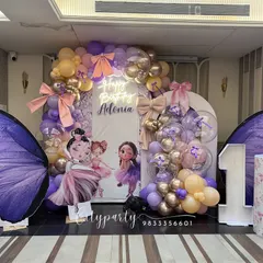 A magical ballerina and butterfly theme for a first birthday party. This elaborate setup features large purple butterfly wing props, ballerina character standees, a light-up marquee number '1', and a balloon arch in pastel purple and gold.