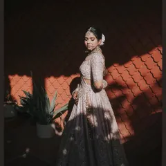 A portrait that tells a story of light and shadow. The bride stands in a sliver of sunlight, her intricate lehenga and delicate features highlighted against a textured, dark background.