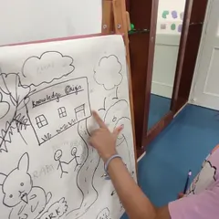 A child points to her drawing of our school, "Knowledge Origin," as she learns about nouns. This activity connects literacy with creative expression, reinforcing learning in a personal way.