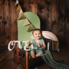 A happy one-year-old boy celebrating his first birthday. This rustic-themed studio setup features a wooden backdrop, a miniature bed, and 'ONE' sign props.