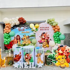 A complete Lion King 'Hakuna Matata' themed birthday setup. It features multiple character cutouts, themed plinths, and an organic balloon garland in jungle-inspired colors.
