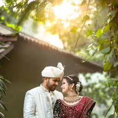 A sun-kissed portrait of the couple in a loving embrace, framed by lush green foliage. The golden light filtering through the trees adds a magical glow to their traditional wedding attire.