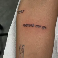 A fine-line Sanskrit script tattoo: 'Yathecchasi तथा kuru', which means 'Do as you wish'. This is paired with a birth year, making it a deeply personal and meaningful piece.
