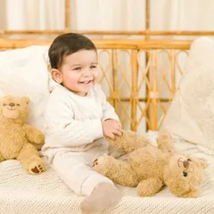 A classic sitter session portrait. This baby's bright, happy smile is captured beautifully in a simple, light-filled studio setting with a rattan bench and some teddy bear friends.
