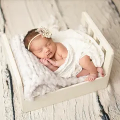 A newborn girl, adorned with a delicate headband, sleeps peacefully in a miniature white bed. I use soft, chunky knit blankets to add texture and warmth to my newborn portraits.