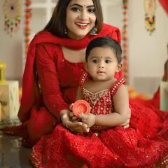 A beautiful mother and daughter portrait from a Diwali mini session. Both are dressed in festive red, with the baby holding a small diya, set against a backdrop of bright, celebratory decorations.