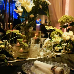 A close-up of a place setting, featuring a textured silver charger plate, a neatly rolled napkin, and a centerpiece of white lilies and floating candles.