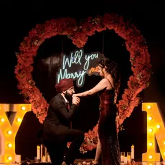 The perfect moment. He gets down on one knee in front of a heart-shaped floral arch and a neon "Will you Marry me?" sign.