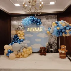 A dreamy sky-themed decoration for baby Avyaan's celebration. This backdrop includes a cloud-painted panel with a personalized light-up name, framed by a blue, yellow, and grey balloon arch and a hot-air balloon prop with a teddy bear.