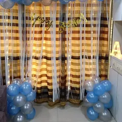 A simple yet beautiful birthday decoration with blue and silver balloon bunches framing a curtain backdrop with fairy lights. This shows how even a small space can be made festive and special with the right balloon arrangement.