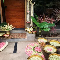 A welcoming entrance for a home-based ceremony, decorated with brass urlis filled with jasmine and rose petals. The traditional rug and hanging floral strings create a warm and inviting atmosphere right from the doorstep.