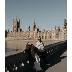 A relaxed moment on a bridge over the Thames, with the historic Houses of Parliament creating a stunning architectural backdrop. The strong sunlight and shadows add a touch of drama to this candid pre-wedding portrait in London.
