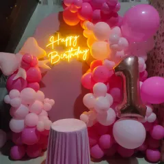 The same pink first birthday setup, now with a 'Happy Birthday' neon sign and a small table for the cake.