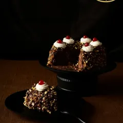 This is our Chocolate Hazelnut Cake, presented with a slice cut out to show the rich interior. It's covered in crunchy, toasted almond pieces and topped with dollops of cream and a single cherry.