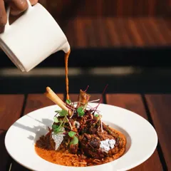 A dramatic shot of lamb shanks being served at a restaurant, using harsh natural sunlight to create strong shadows and a high-end feel.