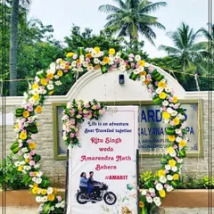 A personalized welcome setup for an outdoor wedding, featuring a custom sign with the couple's names and caricature. The sign is framed by a beautiful semi-circular floral arch, creating a perfect photo opportunity for guests.