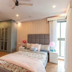 A cozy bedroom for a girl, featuring a cushioned headboard, soft pink paneling, and a white study unit with display shelves for personal treasures.