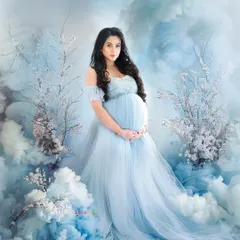 A stunning, wide-angle view of a dreamy maternity shoot. The expecting mother stands in a flowing blue gown, surrounded by a set designed to look like a beautiful sky with clouds and blossoms.