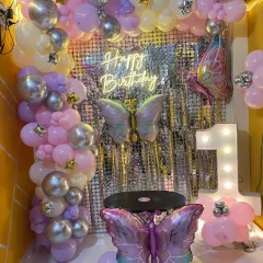 A magical butterfly-themed backdrop for a first birthday. This setup features a silver shimmer wall, a pastel balloon arch in pink and lilac, and large butterfly foil balloons for a whimsical touch.