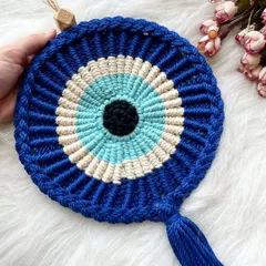 A classic macrame Evil Eye wall hanging, shown here against a soft white background. It features a tassel and a wooden bead for easy hanging.